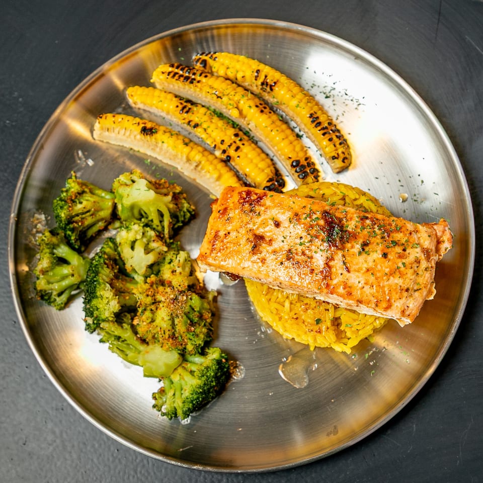 Grilled salmon with yellow rice, roasted broccoli, and corn on the cob on a metal plate.