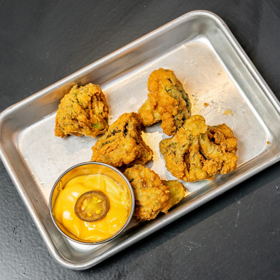 Fried pickles with a side of cheese sauce topped with a jalapeño slice on a metal tray.