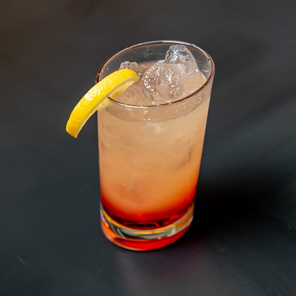 A refreshing cocktail with ice and a lemon wedge, featuring a vibrant red layer at the bottom.
